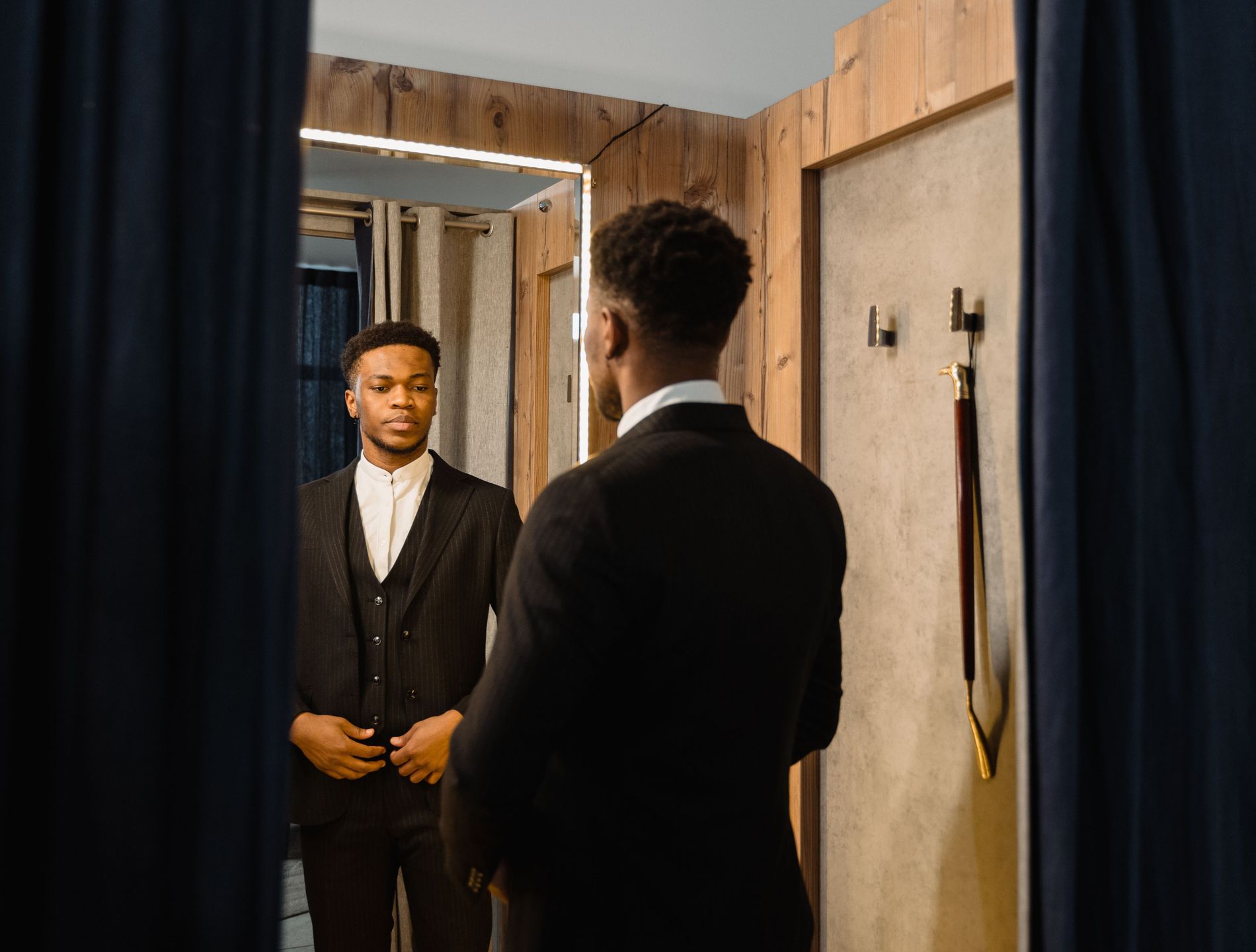 A man in a dark suit checking the fit of his jacket in a mirror, in a private curtained fitting room