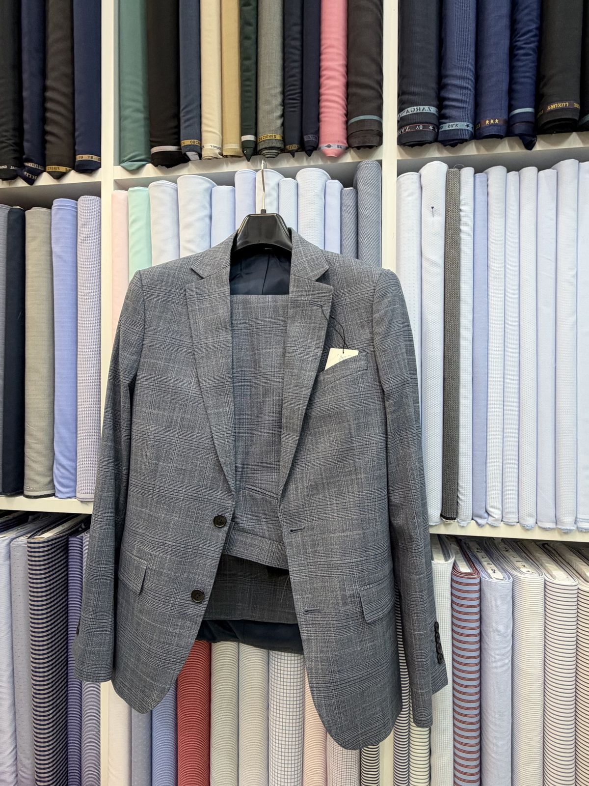 Grey check suit jacket and trousers on a hanger, in front of the shop's cloth shelves
