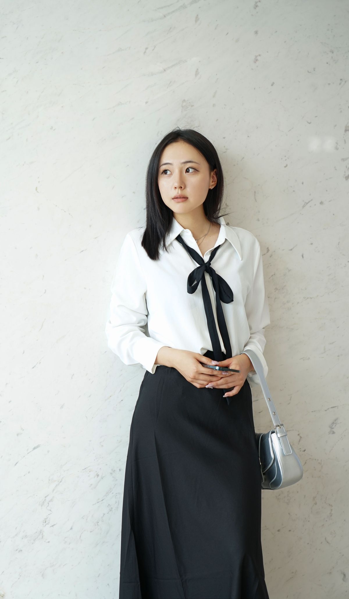 A woman in a white blouse with a black bow and a black midi skirt, against a marble wall