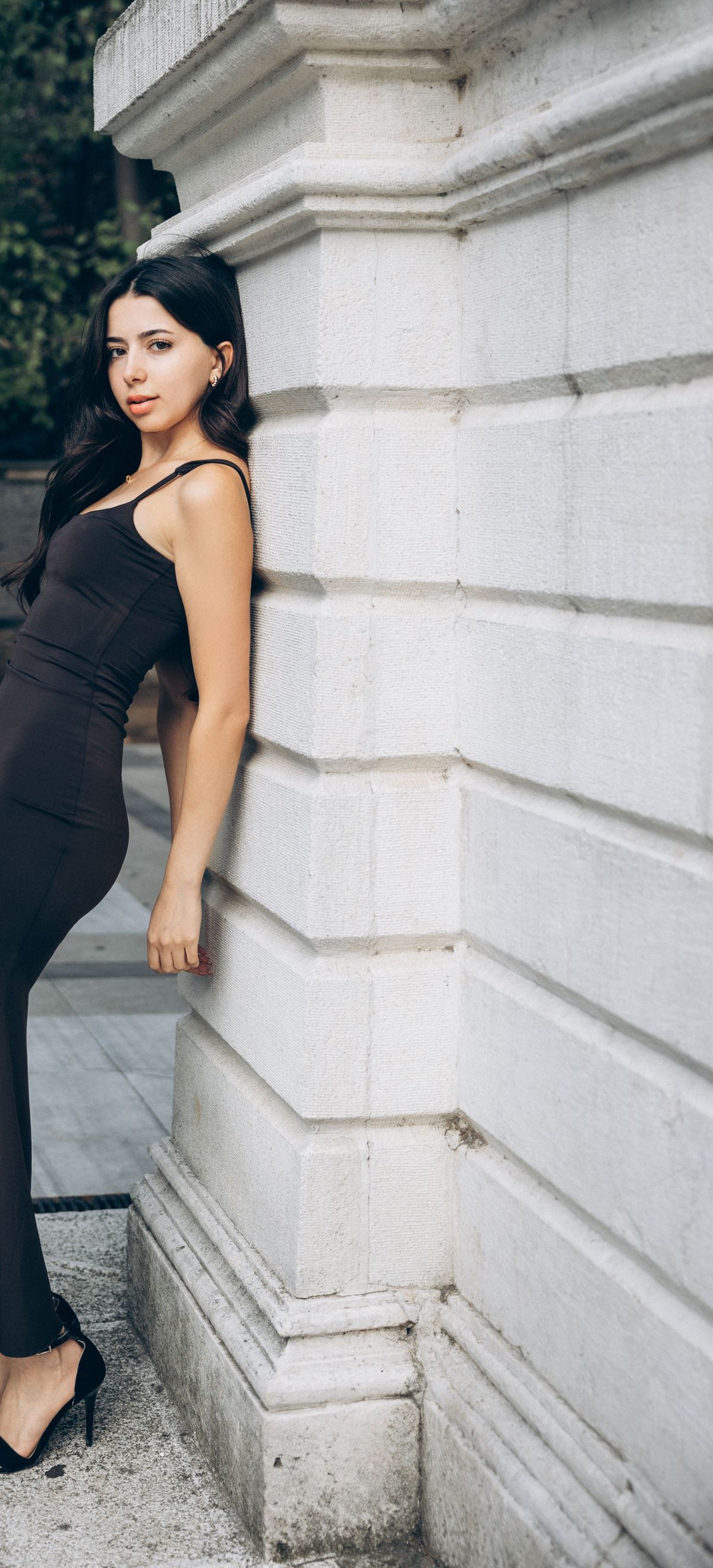 A woman in a fitted black sleeveless dress leaning against a stone building